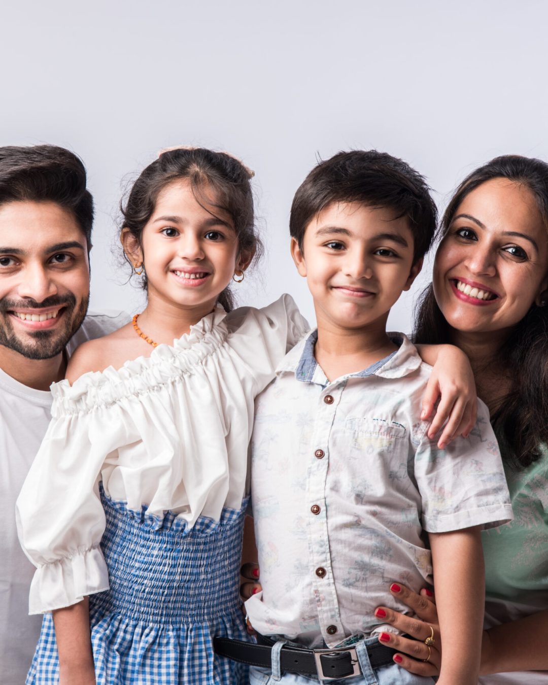 Beautiful Indian young family of four hugging, looking in camera and smiling while standing against white background